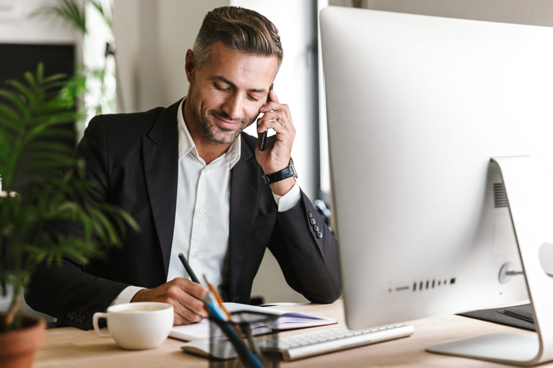 Image of handsome businessman talking on cell phone while workin Image of handsome businessman 30s wearing suit talking on cell phone while working on computer in office