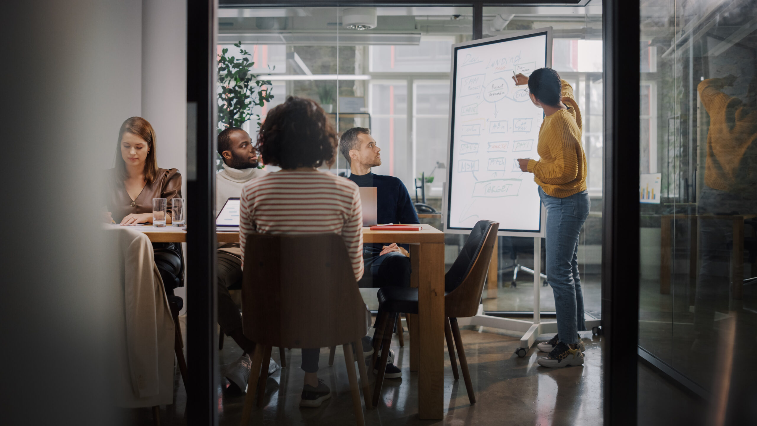 Project Manager Makes a Presentation for a Young Diverse Creativ Project Manager Makes a Presentation for a Young Diverse Creative Team in Meeting Room in an Agency. Colleagues Sit Behind Conference Table and Discuss Business Development, User Interface and Design.
