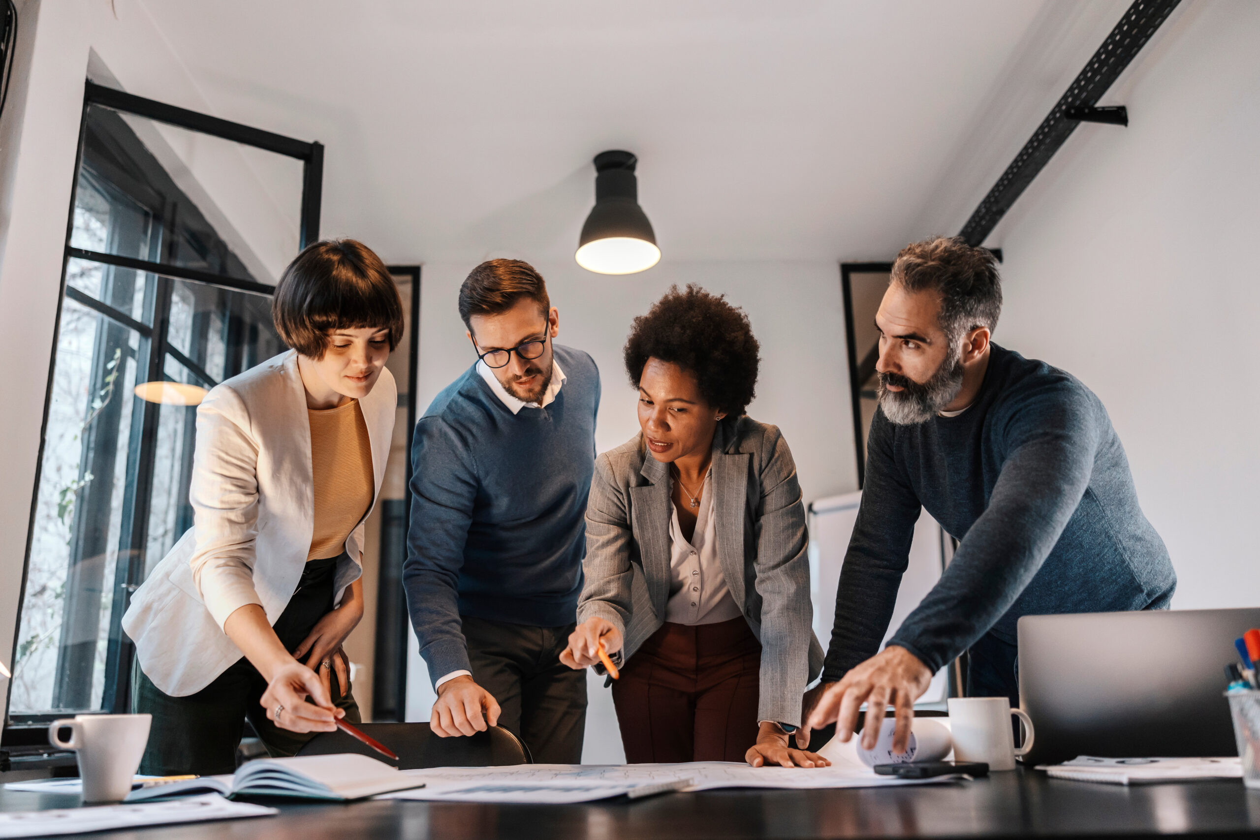 A interracial group of architects is pointing at the blueprints and having briefing about project they working on. A dedicated group of multiracial architects is having meeting at office and looking at blueprints.